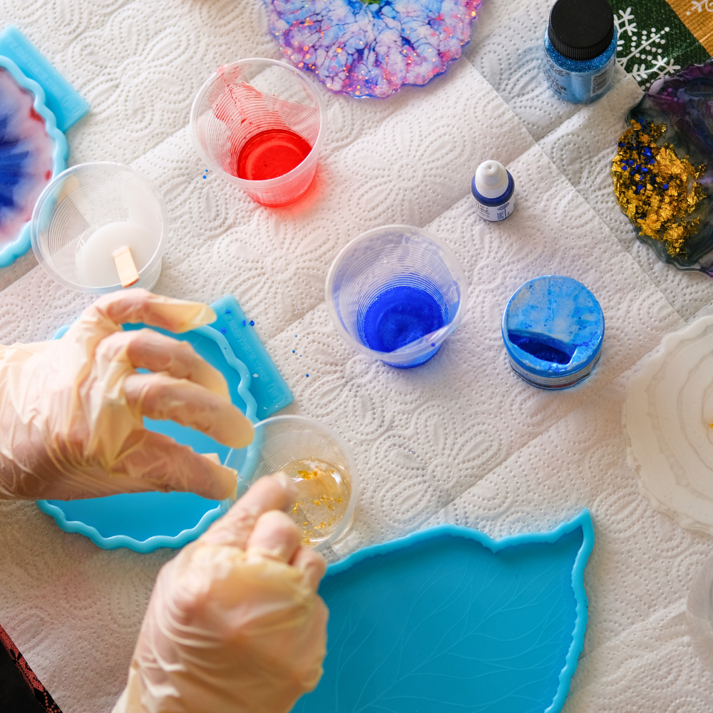 Mãos com luvas preparando artesanato em resina epóxi colorida com moldes e pigmentos.
