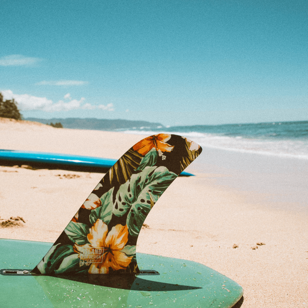 Prancha de surf com quilha floral na areia da praia, mar ao fundo, dia ensolarado